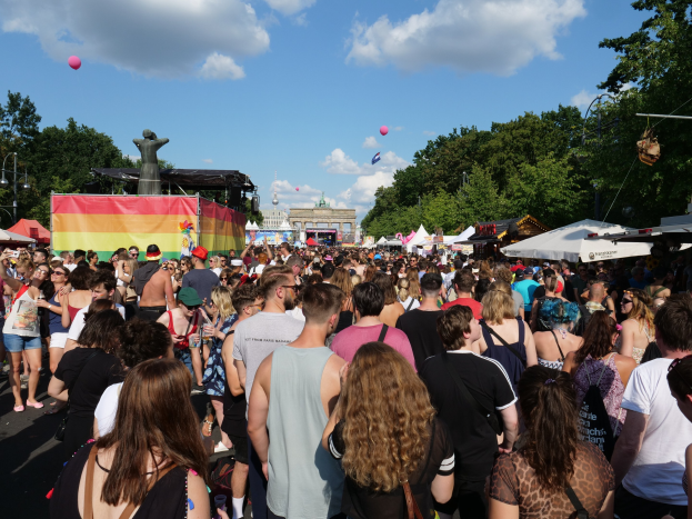 Eine große Menschenmenge geht eine Straße mit Zelten, Bäumen, Pfählen, Lichtern und einer Statue entlang, mit Gebäuden und einem bewölkten Himmel mit Ballons im Hintergrund während des Christopher Street Day Festivals in Berlin.