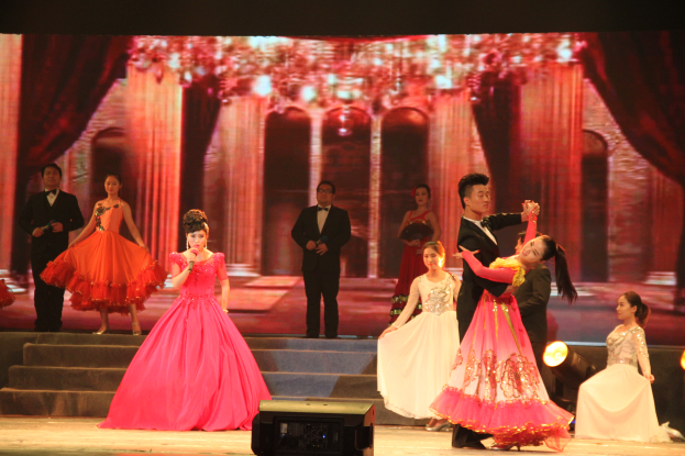 Eine Gruppe von Menschen auf einer Bühne mit Stufen und einem Hintergrundbild, beleuchtet von Scheinwerfern, mit einem Lautsprecher unten, was eine Tanzvorstellung vermuten lässt.