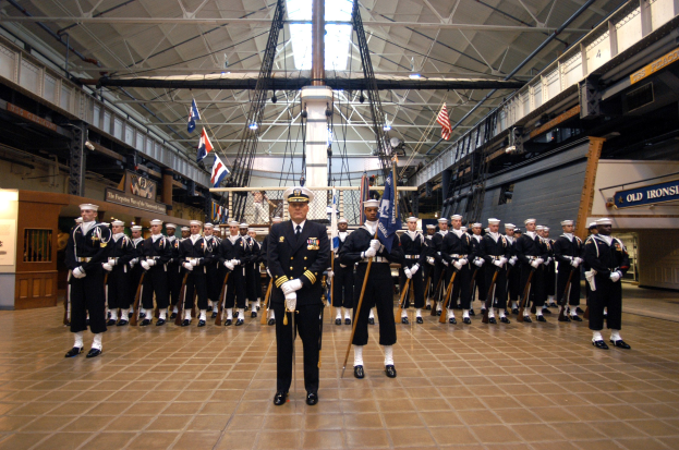 Gruppe uniformierter Männer in einem großen Raum mit Gewehren, Mützen tragend, mit Flaggen und Wandtafeln im Hintergrund.