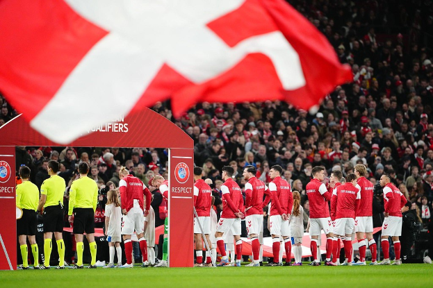 Eine Gruppe von Menschen auf einem Fußballfeld mit einer roten und weißen Flagge im Vordergrund, einem Torbogen mit Text im Hintergrund und einer großen Menge im Stadion.