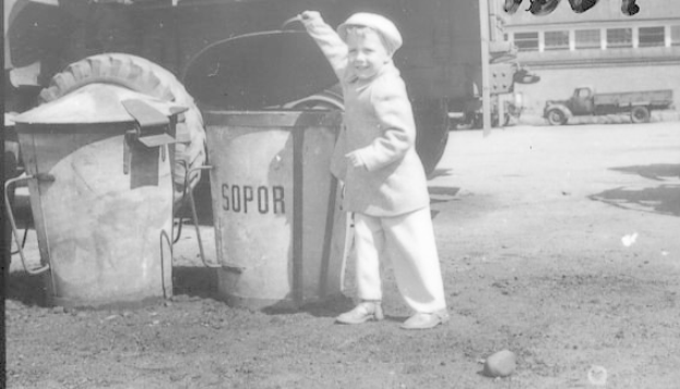Schwarz-weißes Foto eines jungen Jungen, der ein Reifen neben einem Müllcontainer hält, mit Fahrzeugen und einem Gebäude im Hintergrund.