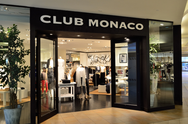 Außenansicht eines Club Monaco Geschäfts in einem Einkaufszentrum mit Glaswänden und -türen, Schaufensterpuppen in Kleidern, Auslagetischen mit Artikeln, einem Topf mit einer Pflanze und Deckenbeleuchtung.