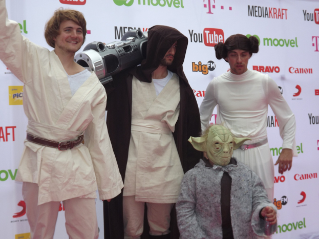 Eine Gruppe von Menschen in Star-Wars-Kostümen posiert auf einem roten Teppich, wobei einer ein Objekt in der Mitte hält und ein Banner mit Text und Logos im Hintergrund zu sehen ist.