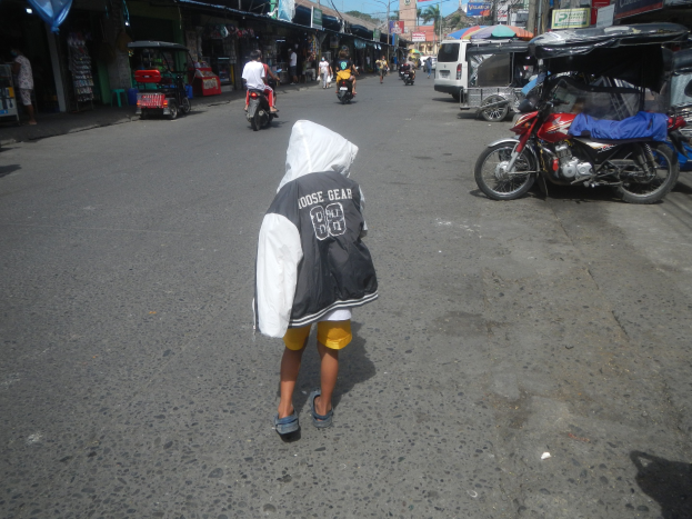 Ein junger Junge geht eine Straße entlang, die von Motorrädern und anderen Fahrzeugen bevölkert ist, flankiert von Gebäuden mit Schildern, unter einem klaren blauen Himmel mit Bäumen und Pfählen im Hintergrund.