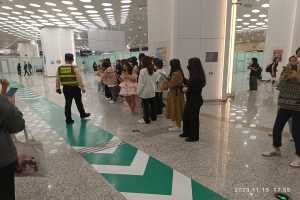 Eine Gruppe von Menschen steht in einer Schlange an einem Flughafen-Sicherheitscheck, einige halten Taschen und Mobiltelefone, mit Säulen im Hintergrund und Lampen an der Decke und einem Wasserzeichen am unteren Bildrand.