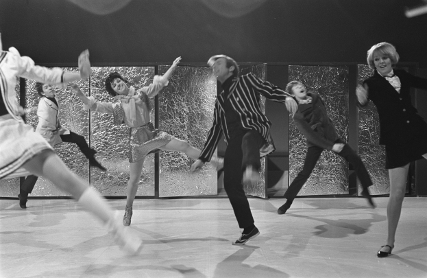 Eine Gruppe von Menschen, die auf einer Bühne vor einer Wand tanzen, alle in Schwarz-Weiß und mit Schuhen.