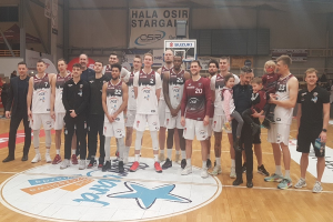 Gruppe von Männern und Kindern auf einem Basketballfeld mit Schildern "Hala Osir Starga", sitzende Zuschauer, ein Basketballkorb, Metallstangen, Deckenlampen und eine Wand im Hintergrund.