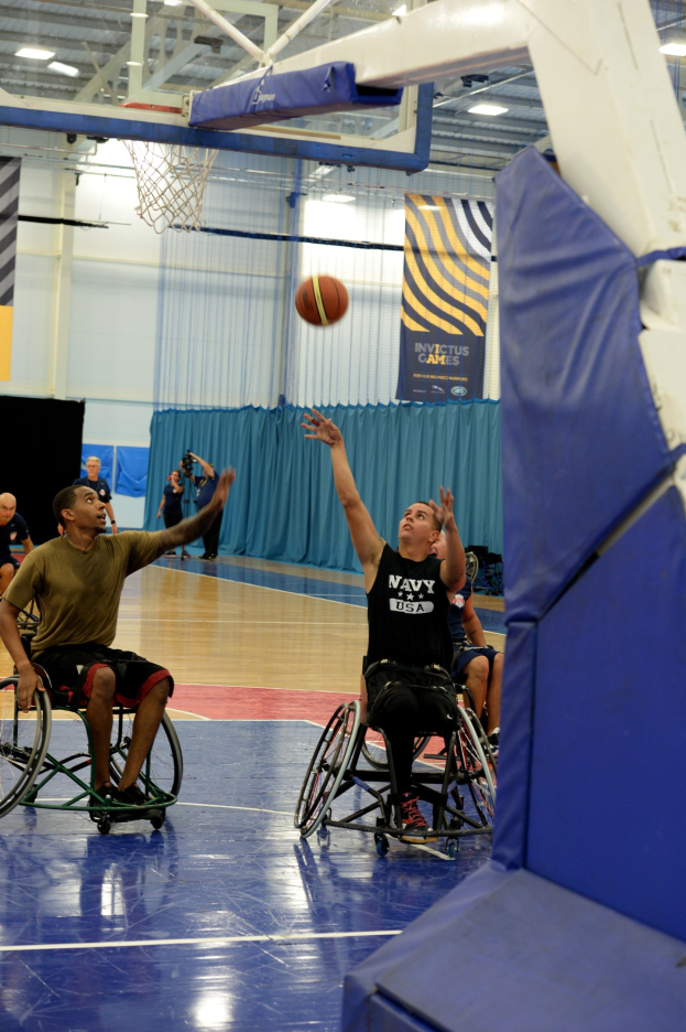 Gruppe von Menschen im Rollstuhl, die Basketball in einer Turnhalle mit Basketballkorb, Vorhängen an der Wand, Bannern mit Text und Deckenlampen spielen.