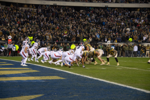 Ein Fußballspiel auf einem Stadionfeld mit Spielern in Sportkleidung und Helmen, umgeben von Zuschauern und Stadionmerkmalen.