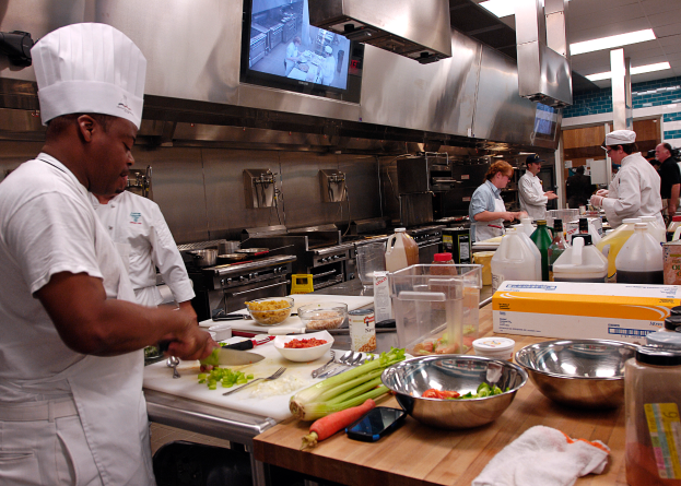 Eine Gruppe von Köchen in einer Küche, die auf einem Schneidbrett Lebensmittel zubereiten, wobei ein Koch im Vordergrund einen weißen Dress und eine Haube trägt, ein Messer hält und von Utensilien, Schüsseln, Gemüse, einem Mobiltelefon, Flaschen und anderen Gegenständen umgeben ist, während im Hintergrund ein Fernseher zu sehen ist.