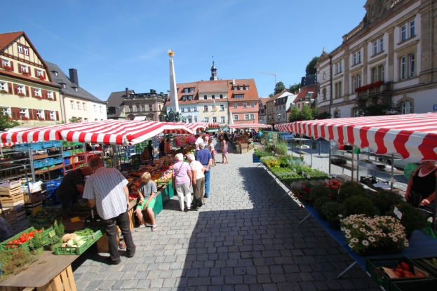Ein belebter Markt im historischen Stadtzentrum von Heidelberg, Deutschland, mit Menschen, die gehen, auf Bänken sitzen und in der Nähe von Zelten, Tischen mit Körben voller Gemüse und Gebäuden mit Fenstern, Bäumen und einem klaren blauen Himmel im Hintergrund.