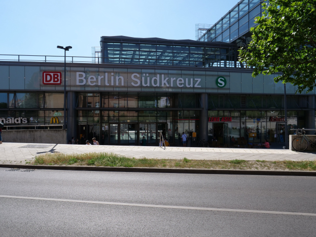 Großes modernes Bahnhofgebäude mit Glaswänden, die Text anzeigen, umgeben von Straßeninfrastruktur, Fahrzeugen, Fußgängern, Fahrrädern, Bäumen und einem klaren Himmel.