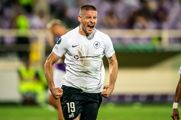 Ein Mann in weißem Hemd und schwarzen Shorts rennt auf einem Fußballfeld und wird von einer Gruppe Menschen umringt, die ein Tor feiern.