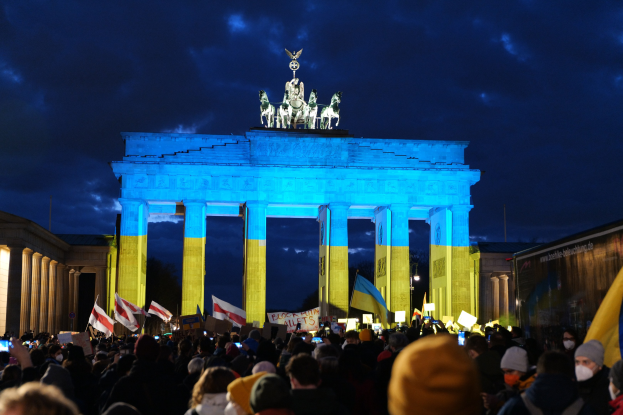 Eine Menge steht vor dem Brandenburger Tor in Berlin, mit Fahnen und Plakaten, einem Banner auf der rechten Seite und den Torstatuen unter einem bewölkten Himmel.