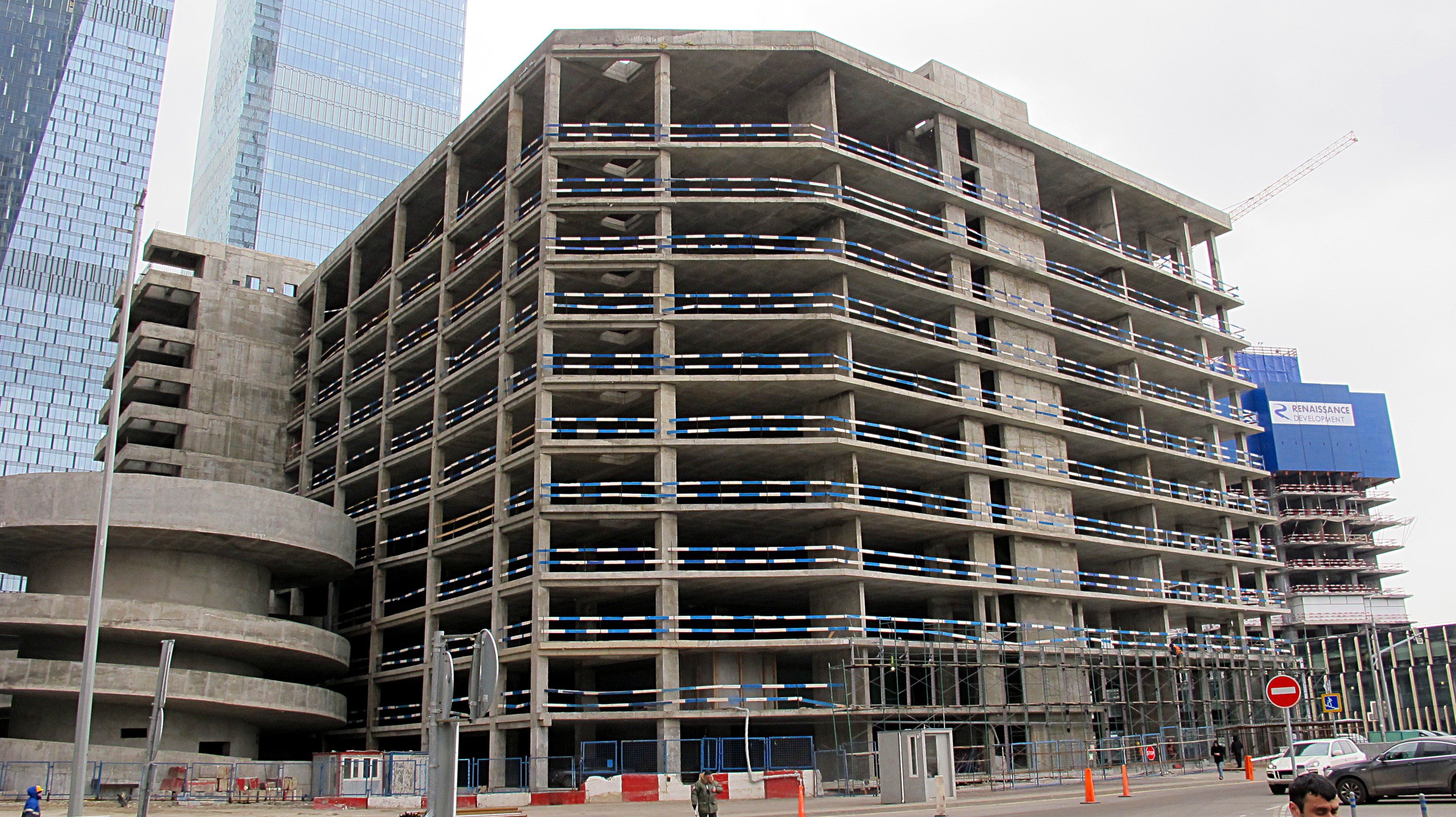 Großer Bau in einer Stadt mit Wolkenkratzern, Fahrzeugen, Fußgängern, Verkehrsinfrastruktur, Kränen und einem klaren Himmel.