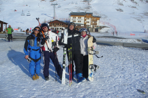 Vier Personen posieren für ein Foto mit Skaterausrüstung und Werkzeugen, mit Gebäuden und anderen Menschen im Hintergrund, vor einer schneebedeckten und vereisten Berglandschaft.
