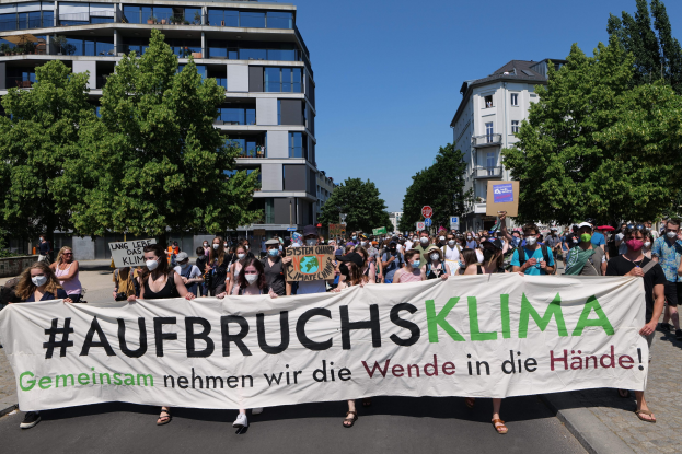 Eine Gruppe von Menschen mit Masken hält ein Transparent mit der Aufschrift "Aufbruchsklima" vor einem von Bäumen und einem klaren Himmel umgebenen Gebäude.