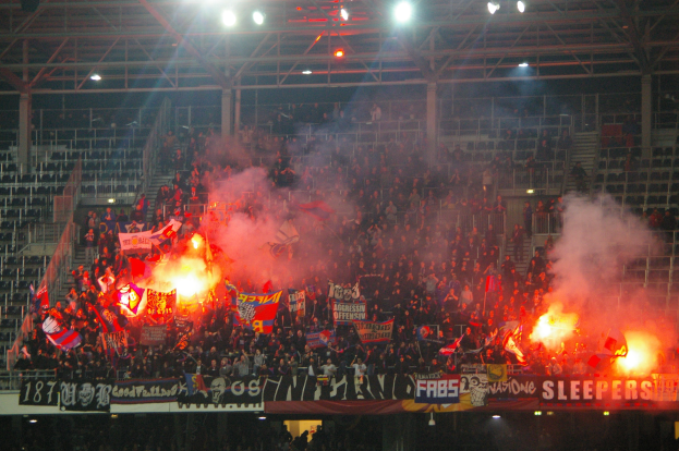 Eine große Menschenmenge in einem Stadion hält Fahnen und Banner, mit Leuchtraketen und Rauch, während Banner, Metallrahmen, Stangen und Deckenleuchten sichtbar sind.