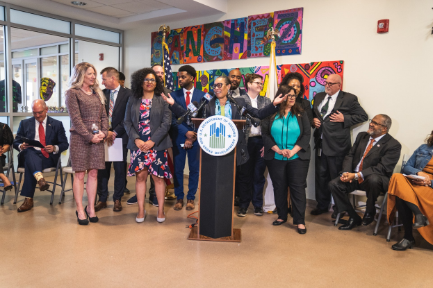 Gruppe von Menschen vor einem Rednerpult mit Mikrofon und einer Tafel mit Text, mit sitzenden und stehenden Zuschauern, Flaggen an der Rückwand und einem Feuerlöscher.