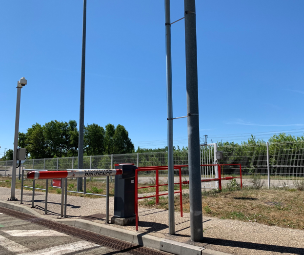Ein Bahnhof mit einem roten und weißen Schild, umgeben von Pfählen, einem Metallzaun, Bäumen, Pflanzen, Gras, einem Straßenpfahl, Drähten und einem bewölkten Himmel, mit einer zentralen Absperrung, um den Zutritt zu verhindern.