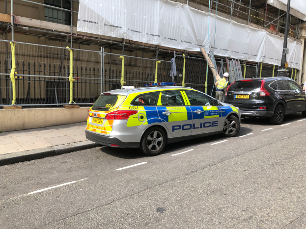 Ein Polizeiwagen steht an der Straße neben einem Gebäude, daneben stehen zwei Helme tragende Personen; im Vordergrund sind ein Geländer und ein Pfahl zu sehen, und im Hintergrund ist ein weißes Tuch über das Gebäude drapiert, was auf eine polizeiliche Untersuchung nach einem Messerangriff in London hindeutet.