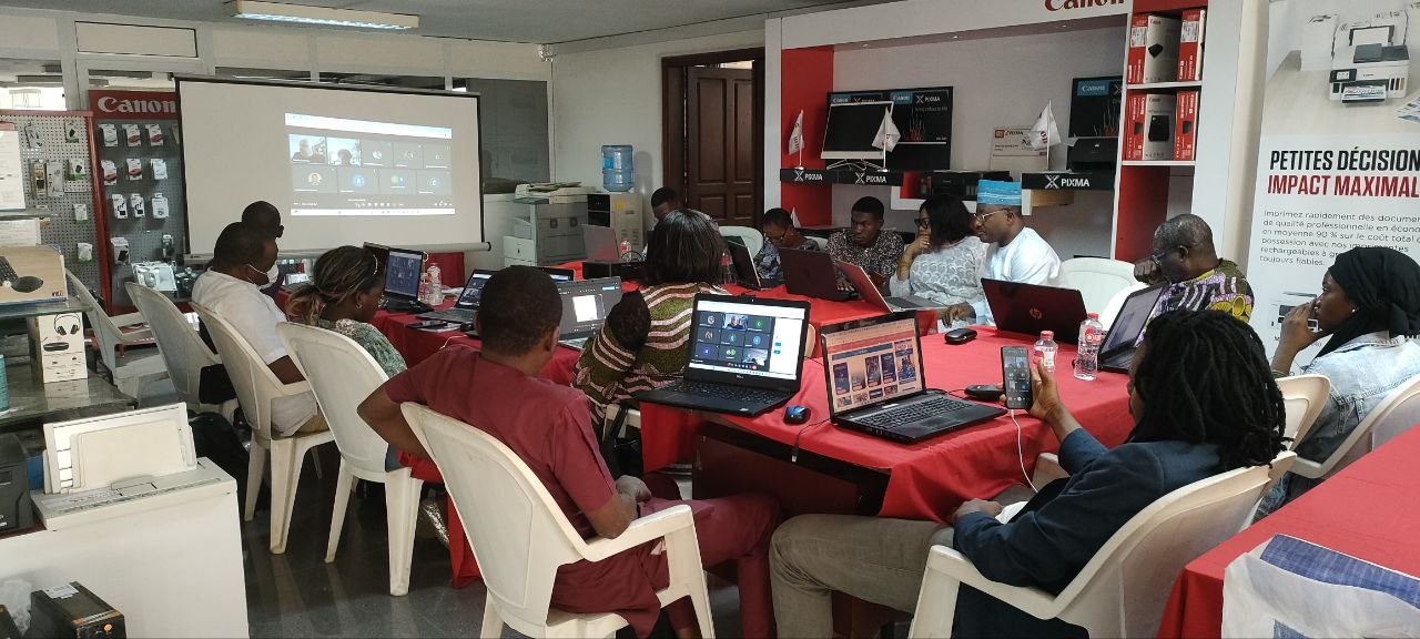 Gruppe von Menschen, die um einen Tisch mit Laptops und anderen Gegenständen während eines digitalen Marketing-Workshops sitzen, mit einem Banner, einem Display und Deckenleuchten im Hintergrund.