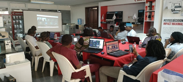 Gruppe von Menschen, die um einen Tisch mit Laptops und anderen Gegenständen während eines digitalen Marketing-Workshops sitzen, mit einem Banner, einem Display und Deckenleuchten im Hintergrund.