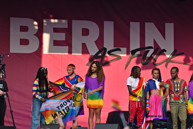 Eine Gruppe von Menschen auf einer Bühne mit einer Frau, die in der Mitte eine Flagge hält, einer Person, die links eine Kamera auf einem Stativ bedient, und einem Banner im Hintergrund mit der Aufschrift "Berlin als Fuck 2019".
