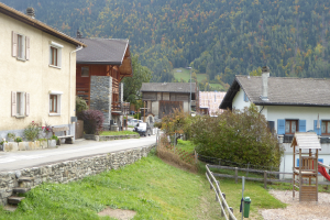 Kleines Dorf mit Häusern, Bäumen, Pflanzen, Gras, einem Holzzaun, einer Rutsche, einem Mülleimer, einer Straße, Fahrzeugen, Pfählen und einem Hügel im Hintergrund.