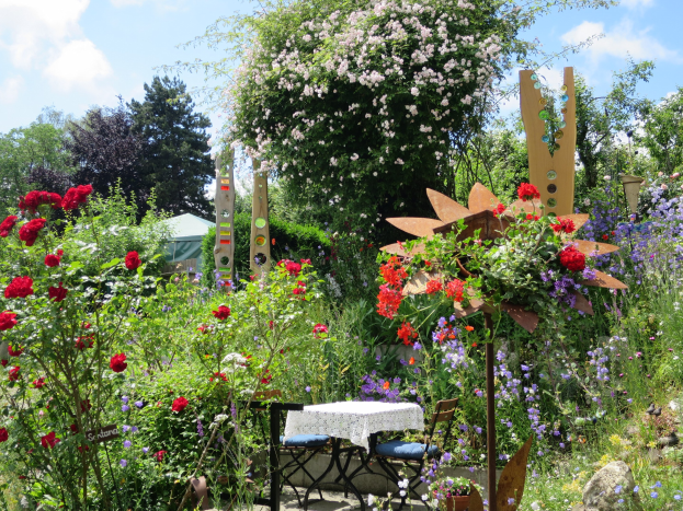 Ein Garten mit bunten Blumen und Pflanzen, der einen Tisch mit einer Decke und Stühlen zeigt, vor Bäumen und einem bewölkten Himmel am Chelsea Flower Show.