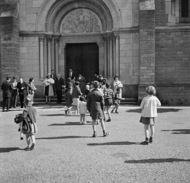 Black and white image of people walking past a building with a door and wall-mounted sculptures.