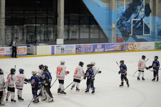 Junge Menschen in Helmen und Eishockey-Ausrüstung spielen Eis-hockey auf einer Indoor-Eisfläche mit Bannern, Säulen und Glaswänden im Hintergrund.