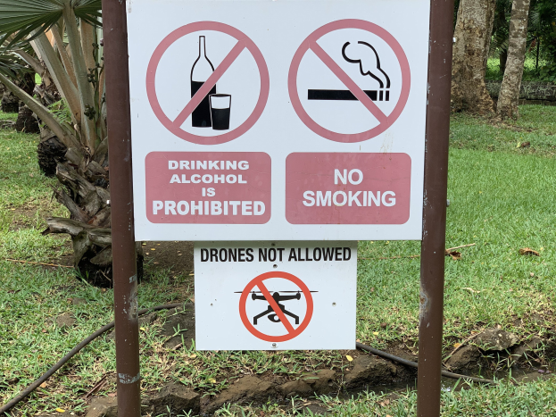 Schild am Straßenrand mit der Aufschrift "Alkoholgenuss ist verboten" und "Rauchen verboten" umgeben von Bäumen und Gras.