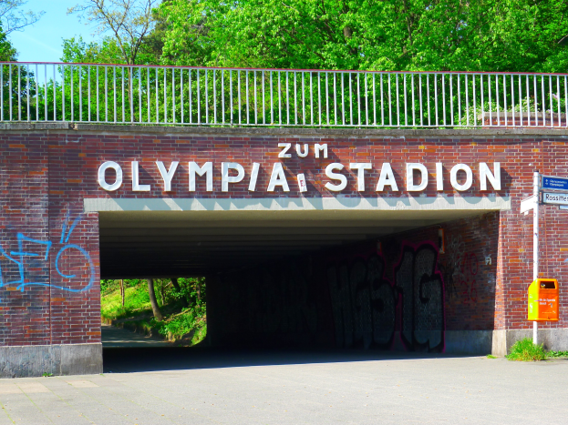 Der Eingang zum Olympiastadion in Berlin, Deutschland, mit einer Brücke mit Text, einem Metallzaun, einer Tafel, einer Box, Pflanzen, Gras, Bäumen und einem bewölkten Himmel.