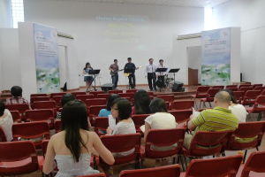 Eine Bühne mit zwei Gitarristen in der Mitte und anderen Musikern in den Ecken, mit Fahnen neben der Bühne und einem Publikum davor mit Papieren und Büchern.