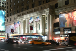 Eine belebte Straße in der Stadt bei Nacht mit fahrenden Fahrzeugen, beleuchtet von Straßenlaternen, mit einem Gebäude mit Fenstern, Pfeilern, Flaggen und beleuchteten Werbetafeln.