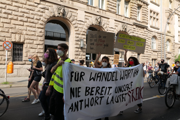 Eine Gruppe von Menschen, die auf einer Stadtstraße demonstrieren, einige halten Schilder und andere fahren Fahrräder, im Hintergrund ein historisches Gebäude mit Rundbogenfenstern, Säulen und Skulpturen.
