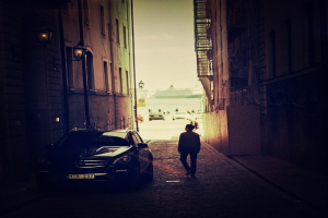 Ein Mann geht auf einem Gehweg neben einem Auto, mit Wänden auf beiden Seiten und einem Pfahl auf der linken Seite; im Hintergrund gibt es Gebäude, ein Schild und mehrere Autos auf der Straße unter dem Himmel.
