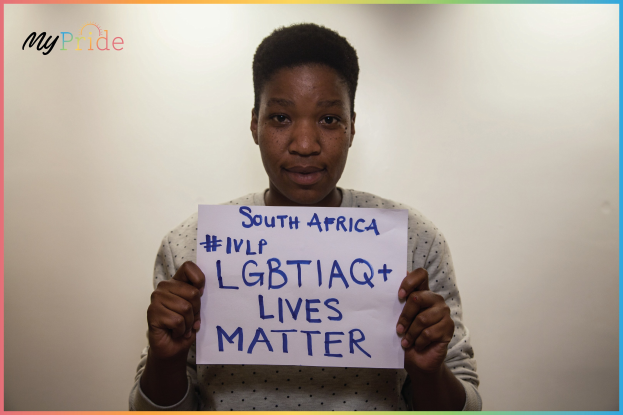 Eine Frau in einem weißen Hemd hält ein Schild hoch, auf dem "LGBTQ+ Lives Matter" steht, vor einer Wand, mit einem entschlossenen Gesichtsausdruck.