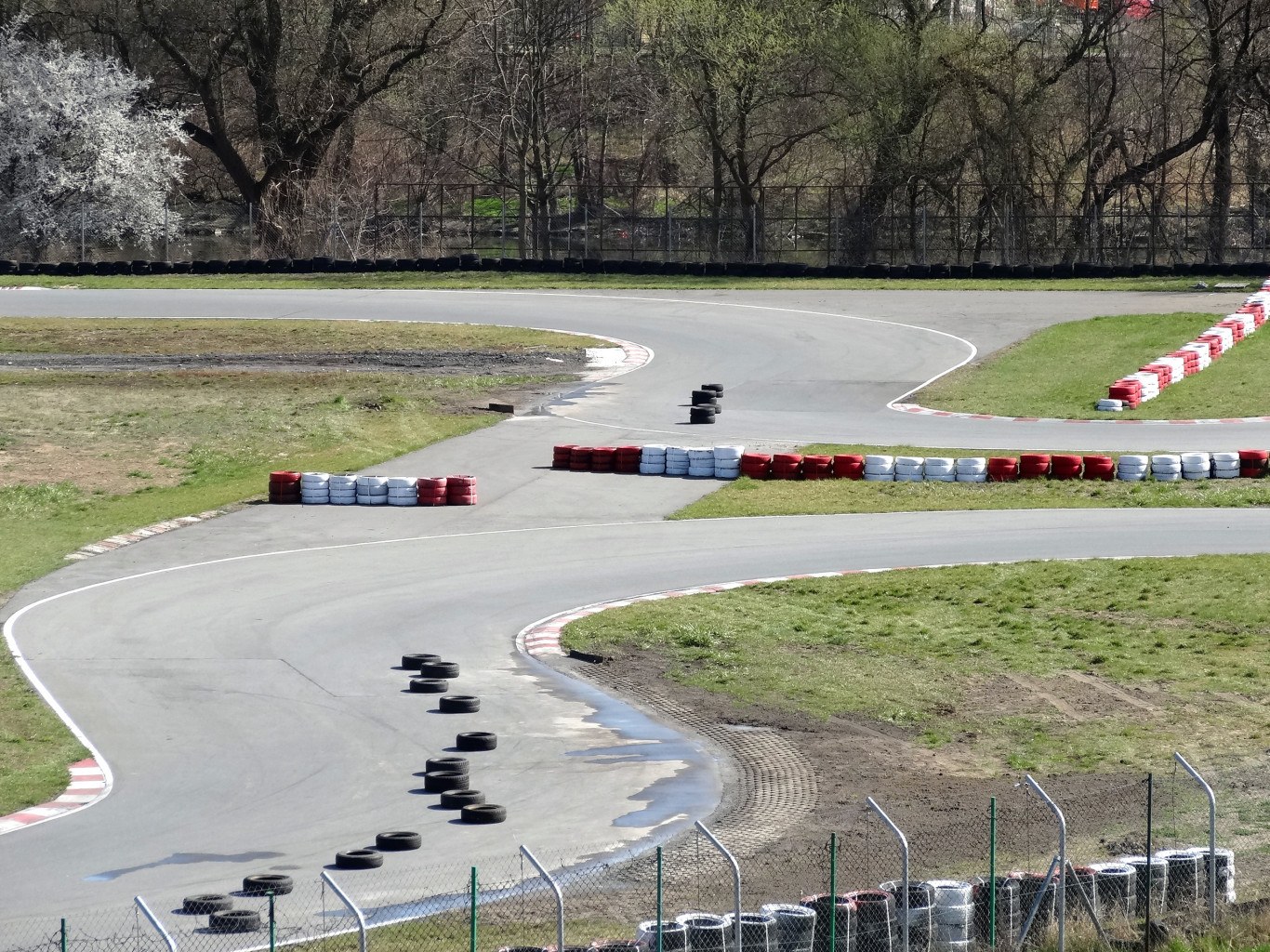 Rennstrecke mit zahlreichen Reifen, die entlang ihrer Ränder angeordnet sind und von Gras und Maschendrahtzaun umgeben ist, mit Bäumen im Hintergrund.