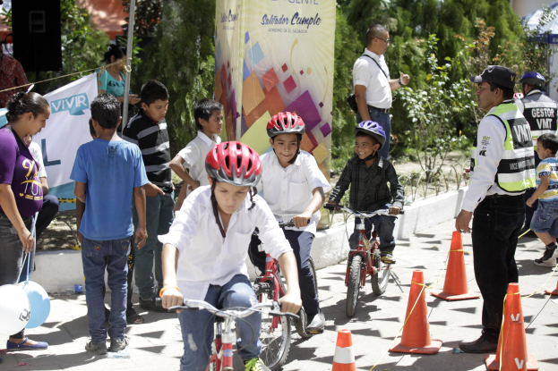 Kinder fahren Fahrräder auf einer Straße mit Verkehrsleitkegeln, einige tragen Helme, andere stehen daneben, mit einer Fahne, Bäumen und Gebäuden im Hintergrund.