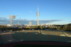 Olympiastadion in Berlin, Deutschland, mit dem Fernsehturm (Fernsehturm) im Hintergrund, umgeben von Bäumen, Gebäuden und beleuchteten Bereichen unter einem bewölktem Himmel.