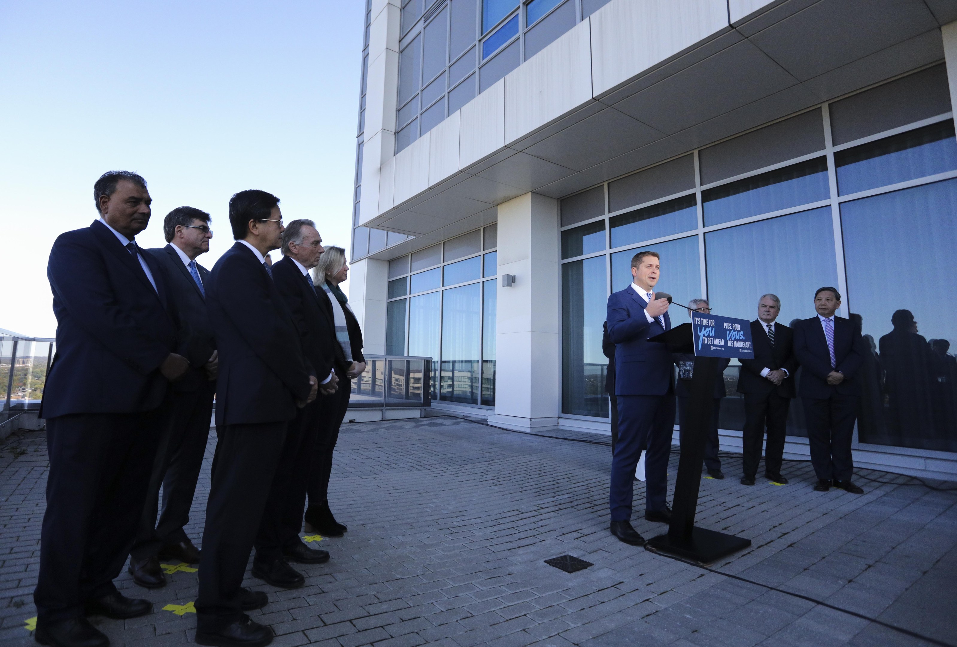 Eine Gruppe von Männern steht vor einem Gebäude mit Glaswänden und Säulen; der Mann in der Mitte hält ein Mikrofon und eine Tafel mit der Aufschrift "Covid-19-Impfstoffausrollung in Russland", mit dem Himmel im Hintergrund.