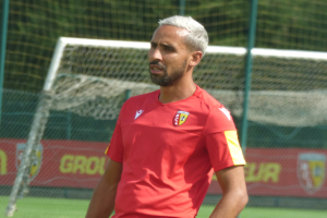 Ein Mann in rotem T-Shirt steht auf einem Fussballfeld und schaut in die Ferne mit einem Netz und einer Flagge mit der Aufschrift "Rajasthan FC" im Hintergrund.