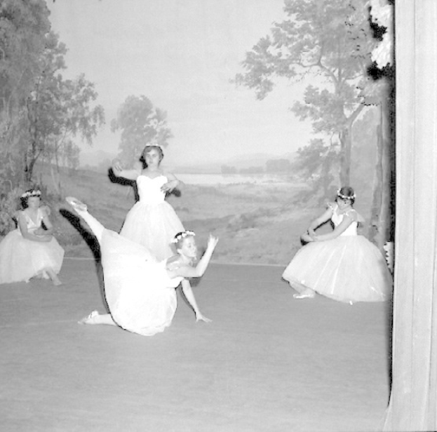 Vier junge Balletttänzerinnen in weißen Kleidern, die auf einer Bühne mit einer gemalten Kulisse von Bäumen und Himmel tanzen, in Schwarz-Weiß.