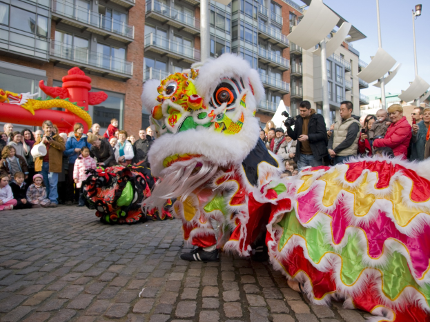 Ein lebendiges chinesisches Neujahrsfest in Amsterdam mit einem Löwen tanzen im Vordergrund, umgeben von einer Menge mit einigen Leuten, die Fotos machen, vor einem Hintergrund von Gebäuden, Laternenmasten und einem klaren blauen Himmel.