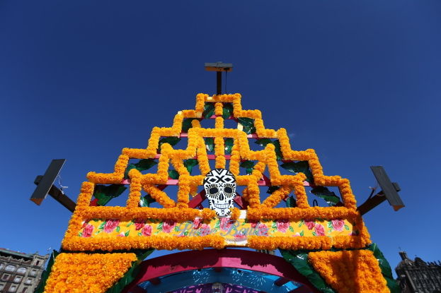Ein Day of the Dead-Bogen in Mexiko-Stadt, geschmückt mit Blüten und einem Schadel, vor einer Kulisse aus Gebäuden unter einem klaren blauen Himmel.