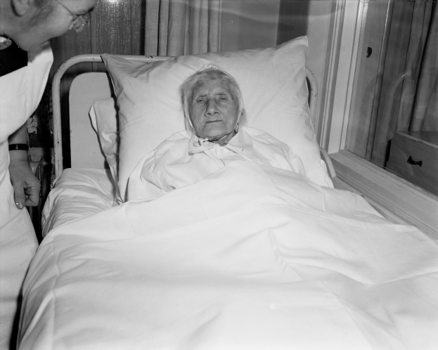 Eine ältere Frau liegt in einem Krankenhausbett mit einer weißen Bettdecke und einem Kissen, während eine Krankenschwester neben ihr steht, mit einem Fenster mit Vorhängen im Hintergrund.