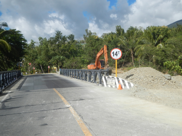 Eine Brücke mit Geländern, ein Schild auf einem Pfahl, ein Bagger, Verkehrskegel, Steine, Bäume, Hügel und ein bewölkter Himmel, mit einer Baustelle und einem Kran auf der Straßenseite.