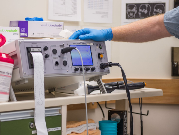 Eine Person in blauen Handschuhen arbeitet an einem medizinischen Gerät auf einem Tisch, der mit verschiedenen Gegenständen übersät ist, mit Papieren an der Wand in einem Krankenhaus.